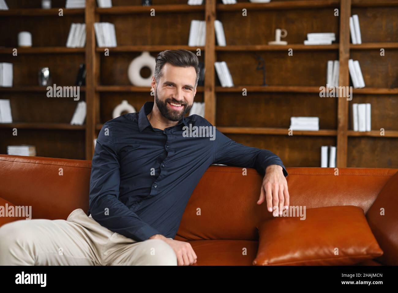 Portrait of a young caucasian man resting on sofa. Handsome positive man with hand on the back of the sofa sitting in living room, daydreaming and relaxing at home Stock Photo