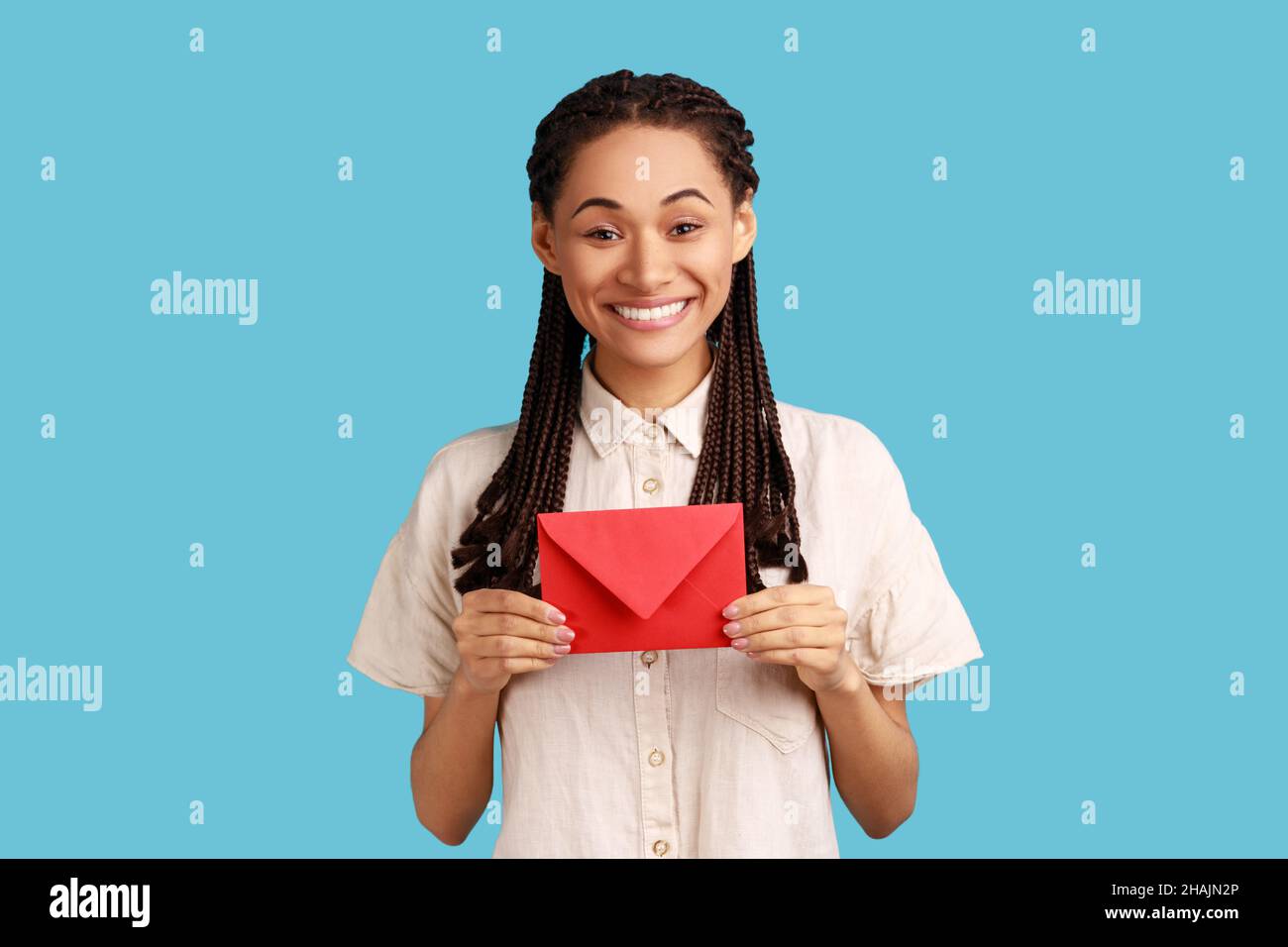 I got love letter on Valentine's day. Happy beautiful woman with black dreadlocks holding letter in red envelope or greeting card and smiling joyfully. Indoor studio shot isolated on blue background. Stock Photo