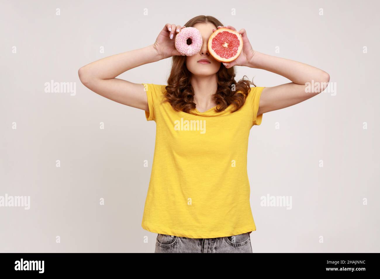 Sugary dessert vs fruits. Portrait of playful brown haired woman in yellow T-shirt covering eyes with fresh grapefruit and sweet doughnut, junk food. Indoor studio shot isolated on gray background. Stock Photo