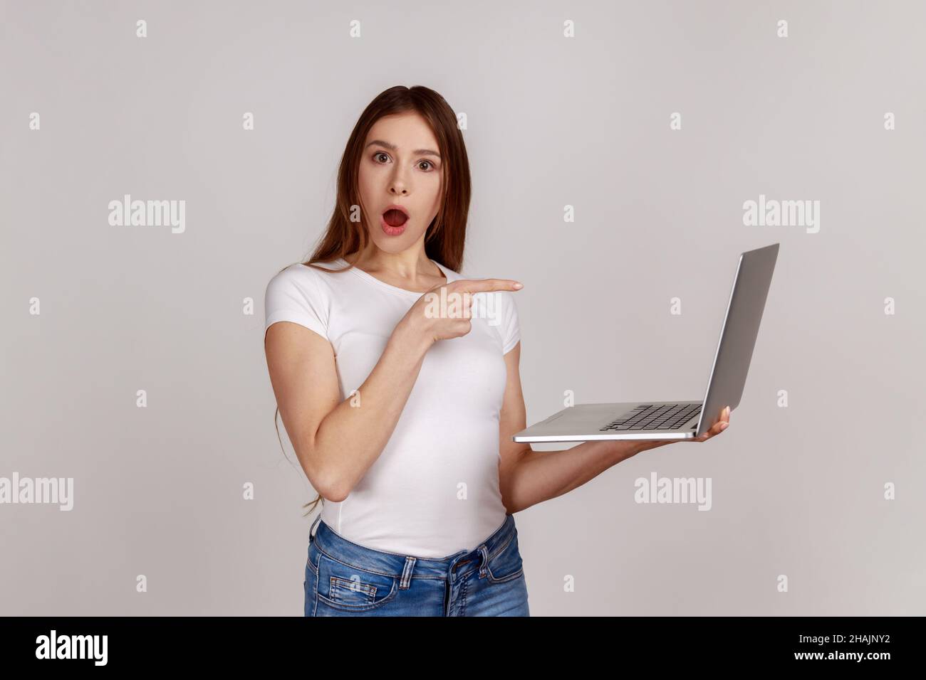 Portrait of amazed emotional woman pointing laptop screen and looking at camera with shocked expression, open mouth, wearing white T-shirt. Indoor studio shot isolated on gray background. Stock Photo