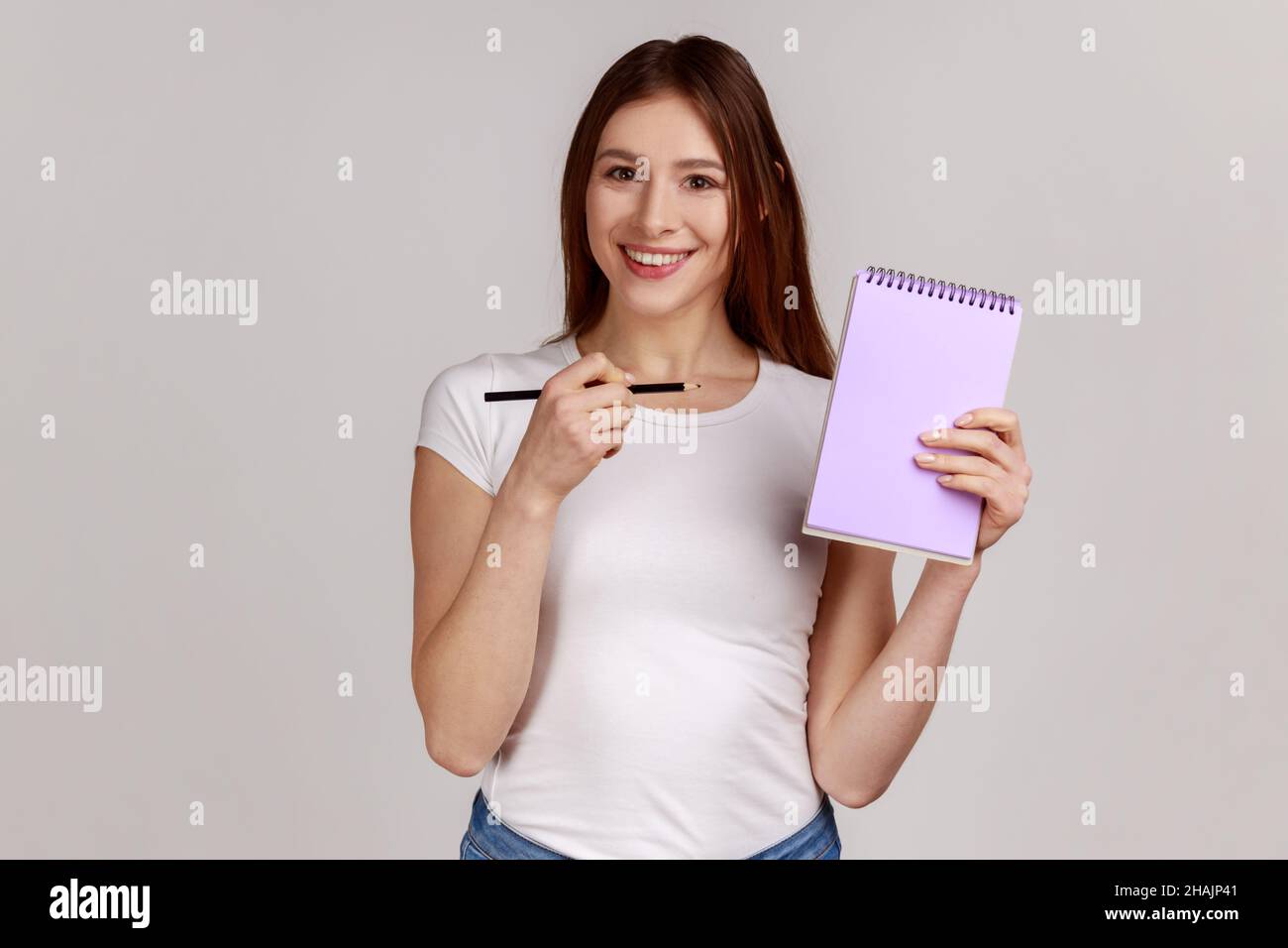 Positive smiling happy woman pointing at paper notebook with blank space for commercial text, advertising area, wearing white T-shirt. Indoor studio shot isolated on gray background. Stock Photo