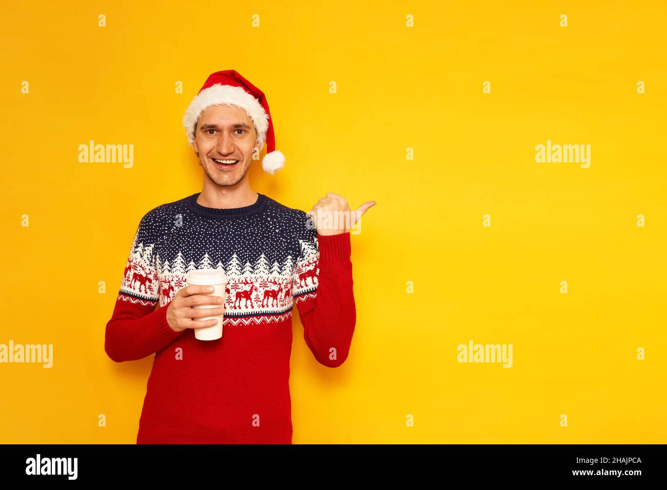 excited man in Christmas clothes sweater with reindeer red Santa Claus hat holds paper cup coffee to go his finger at an empty area for advertising isolated on yellow background with space for text. High quality photo Stock Photo