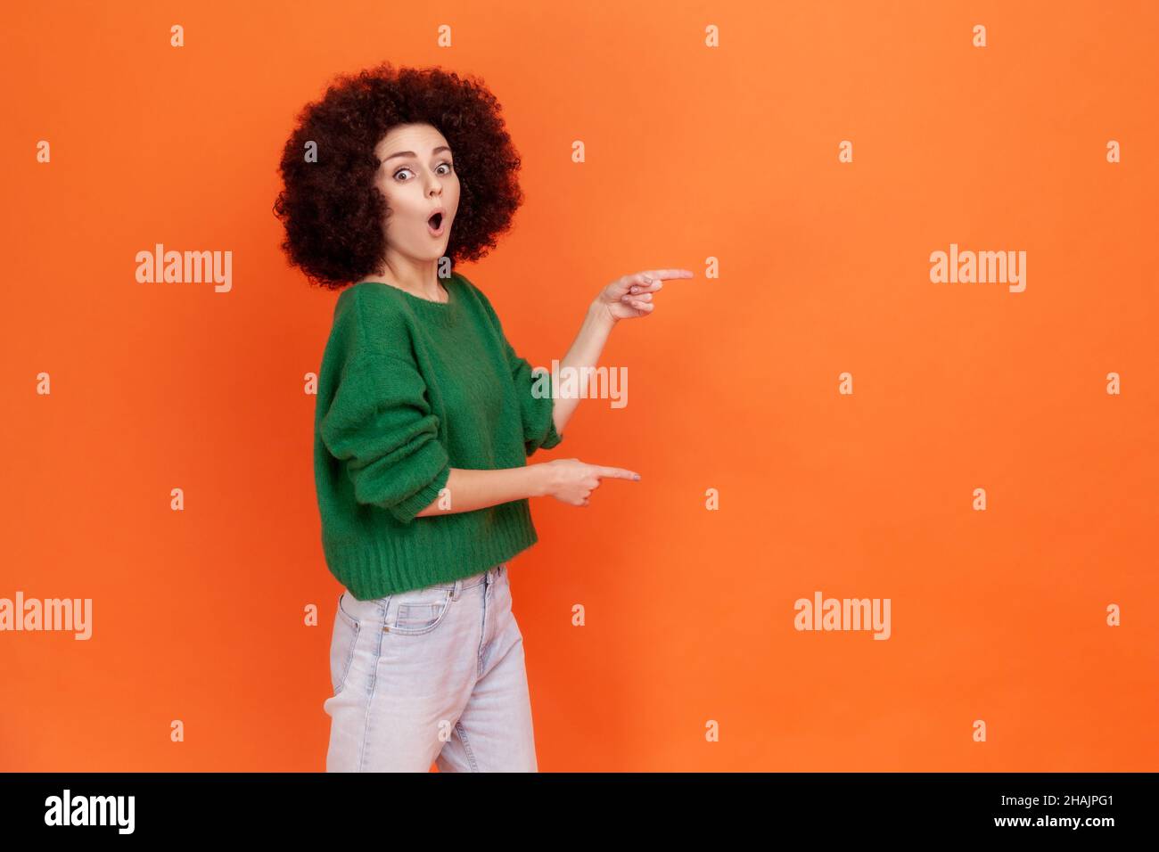 Astonished woman with Afro hairstyle in green sweater pointing aside with finger, having surprised facial expression, keeps mouth open, copy space. Indoor studio shot isolated on orange background. Stock Photo