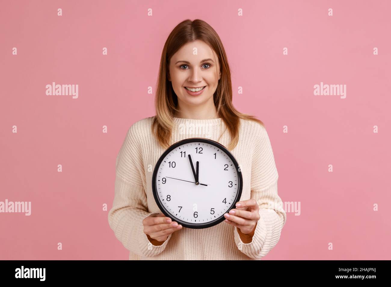 Portrait of happy positive blond woman holding big wall clock, looking at camera with pleasant smile, time to go, wearing white sweater. Indoor studio shot isolated on pink background. Stock Photo