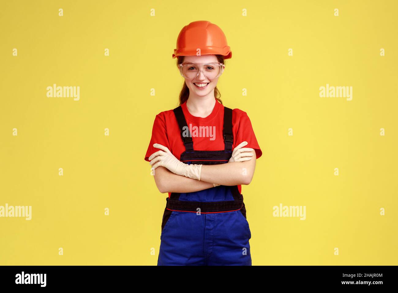 Portrait of positive worker woman standing with crossed arms and looking at camera with confident expression, wearing overalls and helmet. Indoor studio shot isolated on yellow background. Stock Photo