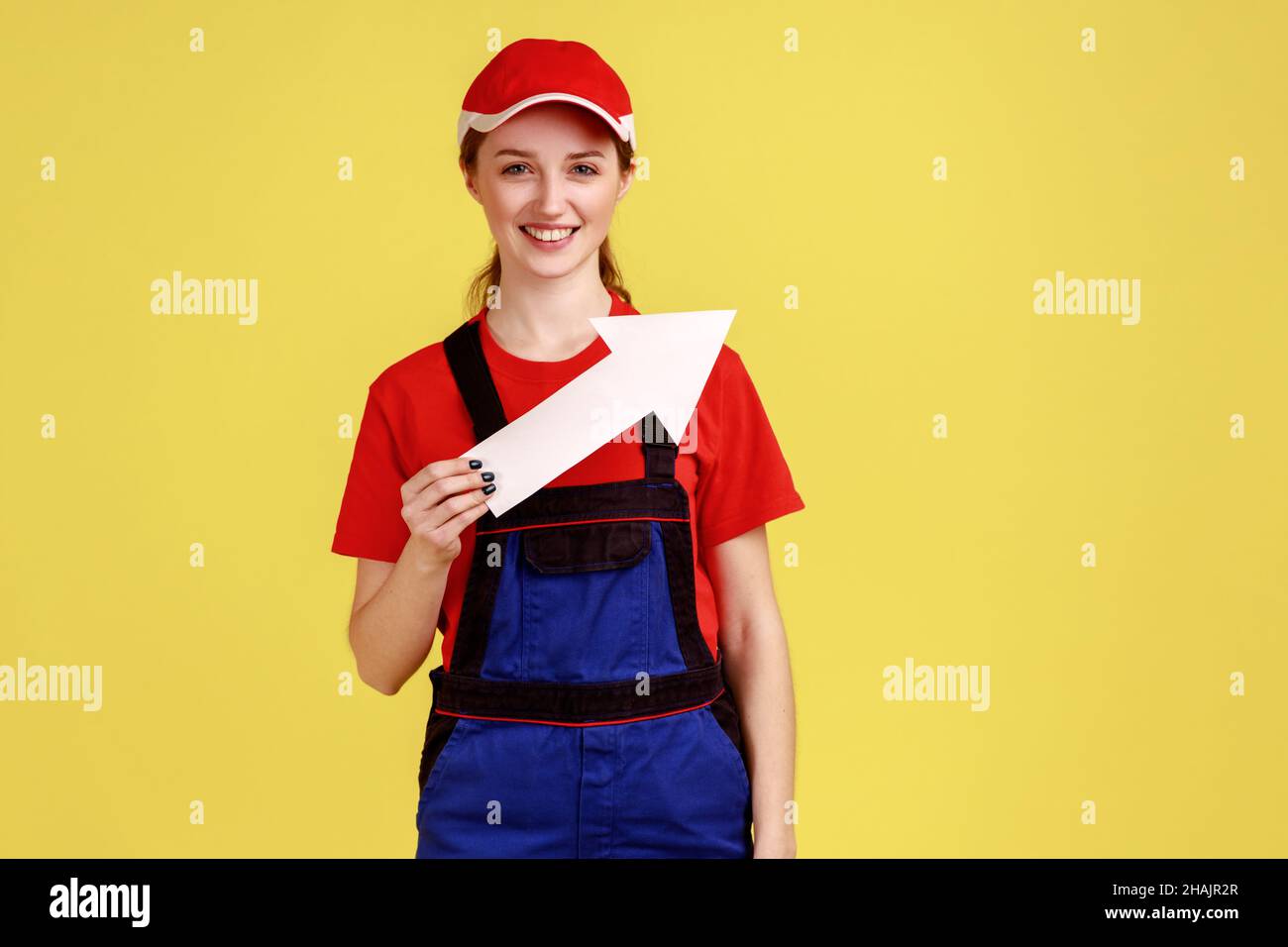 Portrait of smiling positive craftswoman standing with white arrow in hands indicating aside, looking at camera, wearing overalls and red cap. Indoor studio shot isolated on yellow background. Stock Photo