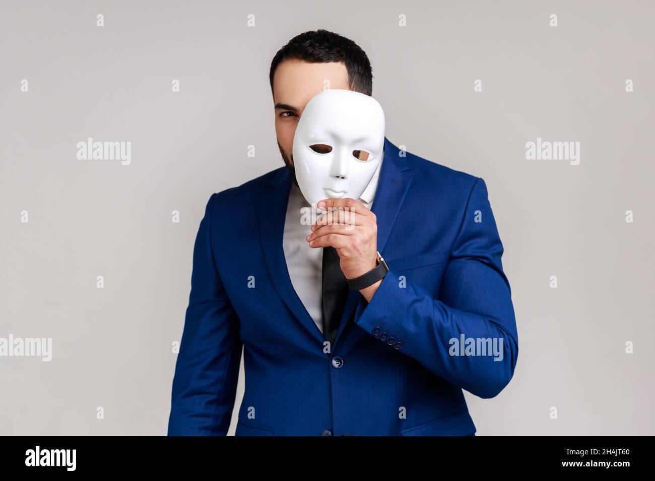 Bearded man holding white mask, covering face, standing with serious expression, multiple personality disorder, wearing official style suit. Indoor studio shot isolated on gray background. Stock Photo