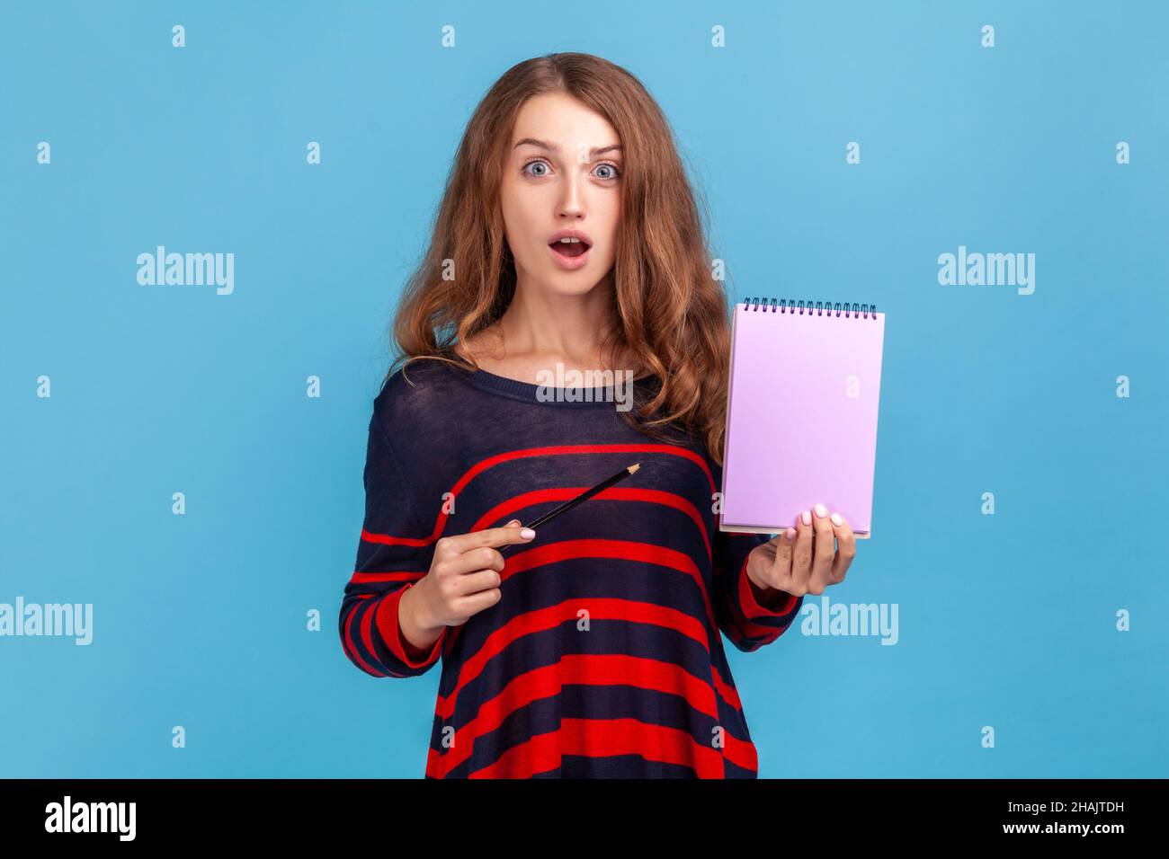 Shocked woman wearing striped casual style sweater, showing blank notebook, advertising area on paper, copy space for to-do list, idea. Indoor studio shot isolated on blue background. Stock Photo
