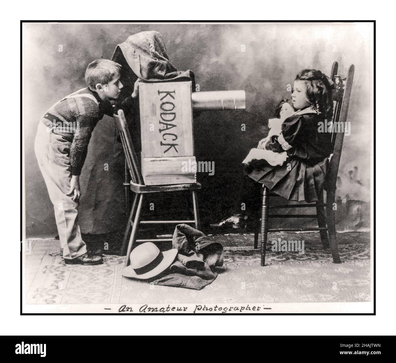Vintage 1900s charming lifestyle photograph 'An amateur photographer' early humourous studio lifestyle photograph featuring deliberate typo miss-spelt Kodak Photograph shows boy with homemade 'Kodack' camera pretending to photograph little girl with doll. Spencer, George W., photographer Created / Published c1907. Children--1900-1910 Photographers--1900-1910 -  Photography--1900-1910 Photographic prints--1900-1910. Stock Photo