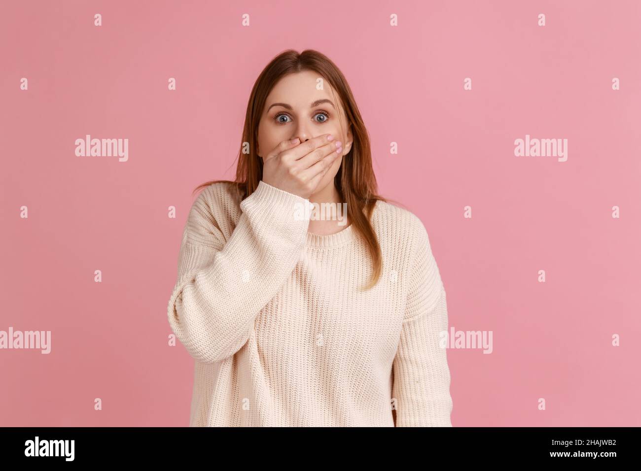 Portrait of shocked blond woman put hand on mouth and looking with fear in her eyes, keeping terrible secret, wearing white sweater. Indoor studio shot isolated on pink background. Stock Photo