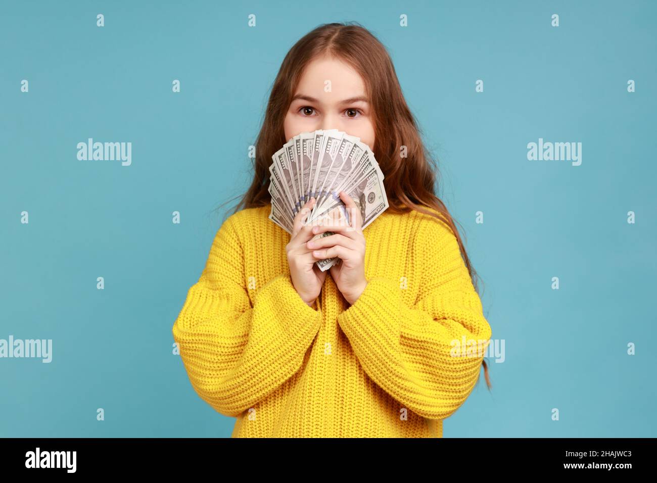 Portrait of little girl hiding half face with money, peeking out of dollar bills, looking at camera, wearing yellow casual style sweater. Indoor studio shot isolated on blue background. Stock Photo