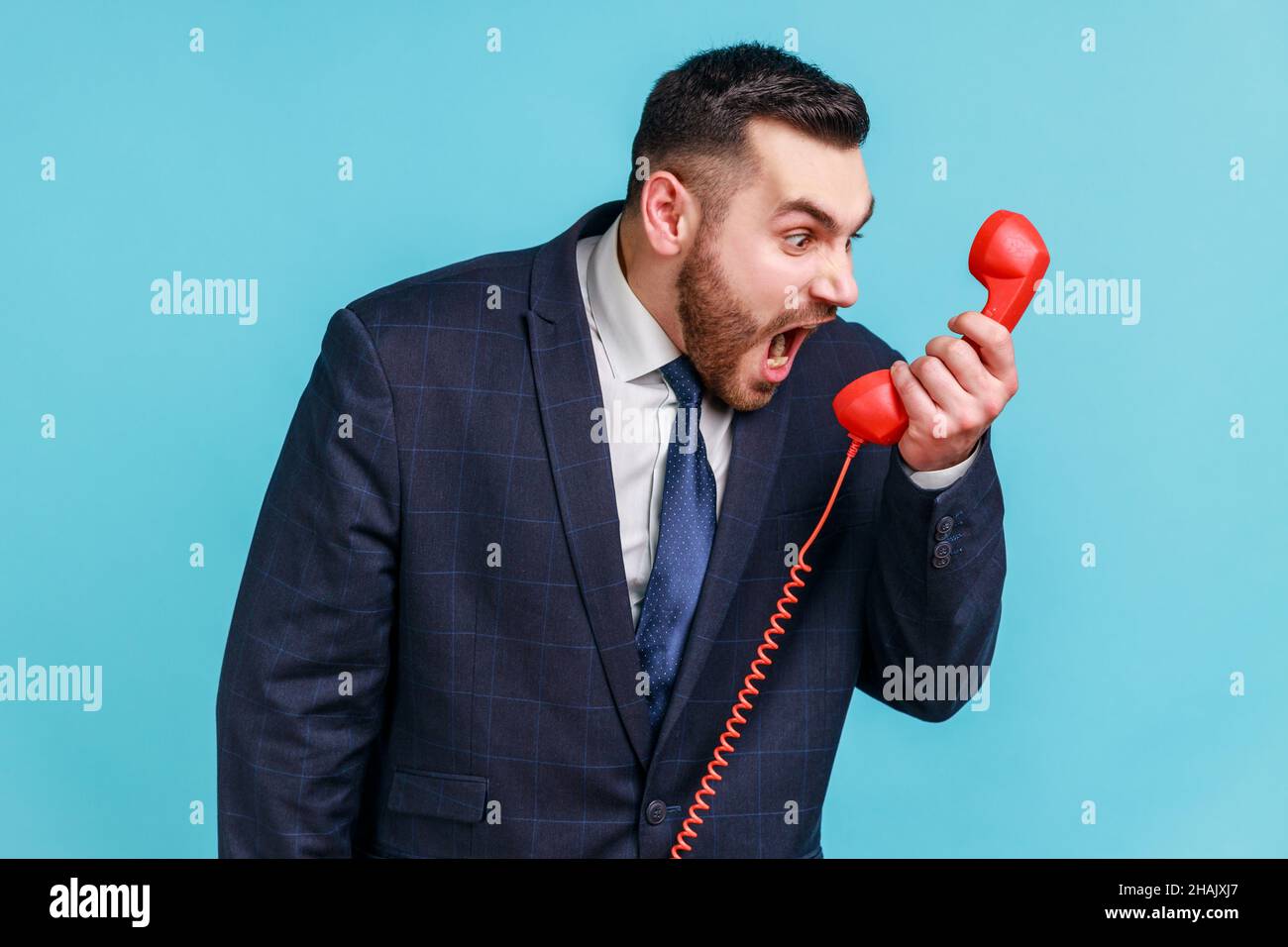 Angry nervous bearded man wearing official style suit screaming and yelling talking retro landline phone, complaining on connection quality. Indoor studio shot isolated on blue background. Stock Photo