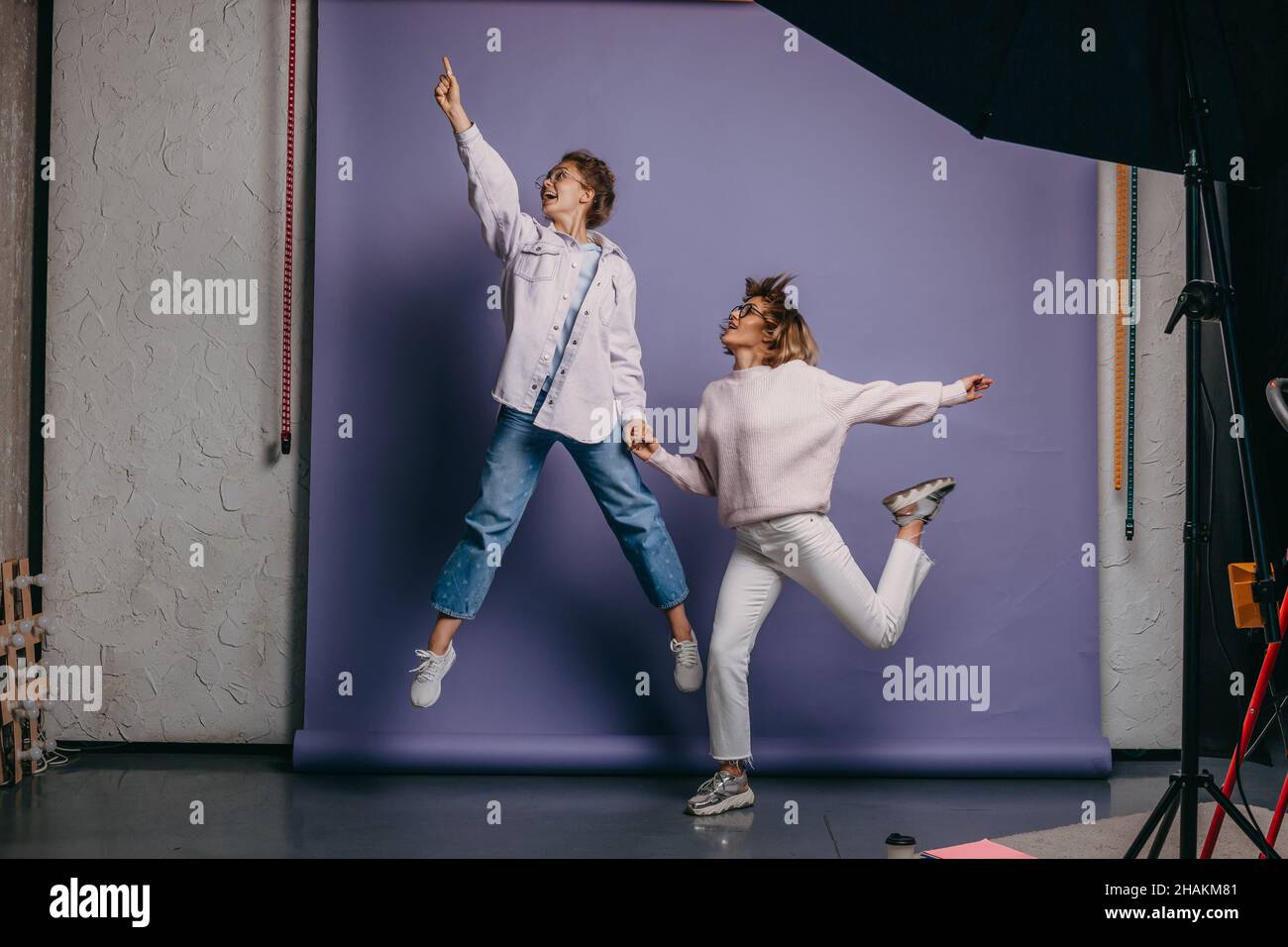 Two women in casual wears jumping on isolated purple background. Hand in hand. Friendship. Stock Photo