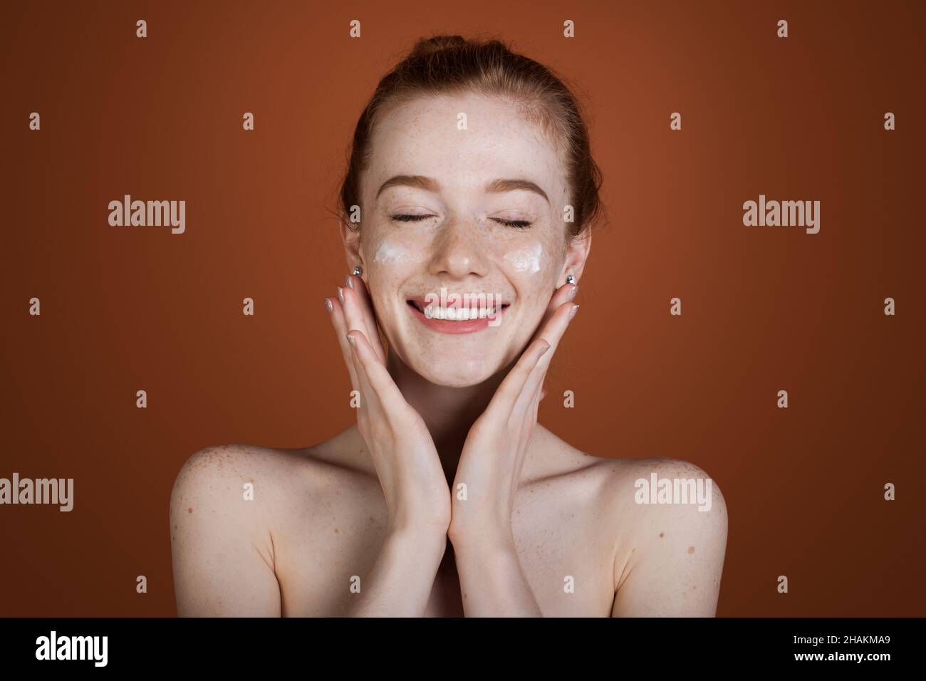Caucasian freckled woman applying cosmetic cream on her face isolated over brown background. Smiling redhead woman. Stock Photo