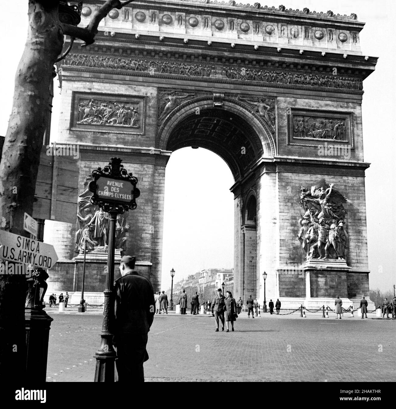 Paris Arc de Triomphe at Place de l Etoile and Champs-Elysees with no vehicles, 1945. The iconic arch takes up most of the square image. Place de l Etoile looks more like a pedestrian plaza then a motorway as no vehicles are in sight. The camera position is where the Avenue des Champs-Elysees ends at the Place de l Etoile. American service personal are visible both in the near ground and middle ground of the image. A stenciled direction sign posted on a tree reads SIMCA US Army ORD. Stock Photo