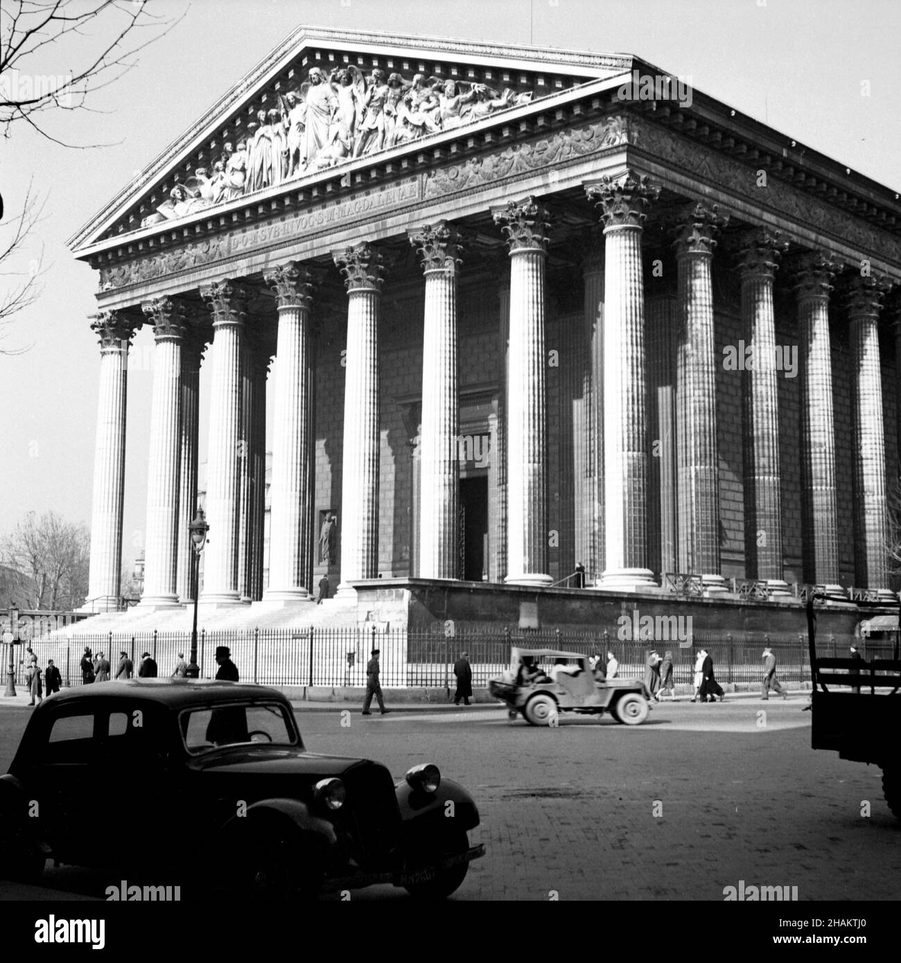 Paris La Madeleine near the end of WWII, 1945. A view of the Neo-classical church which opened in 1842. The camera position is across the road in the southeast corner and depicts the full columned southwest façade and some of the southeast façade. In the road are one military truck, one military jeep, and one parked passenger car. About two dozen pedestrians are in view Stock Photo