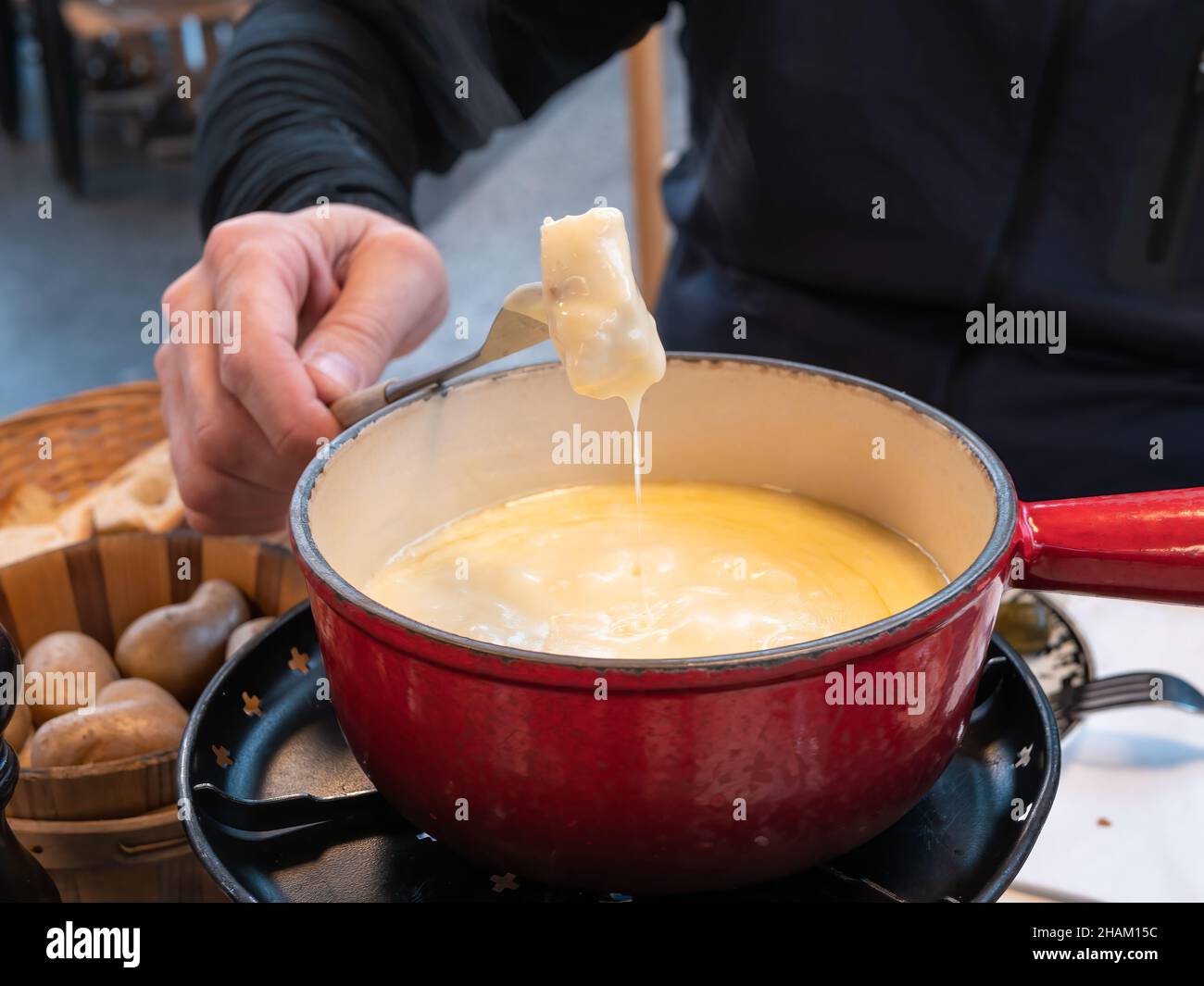A man’s hand holds on a fork a piece of bread that has been dipped in a cheese fondue pot Stock Photo