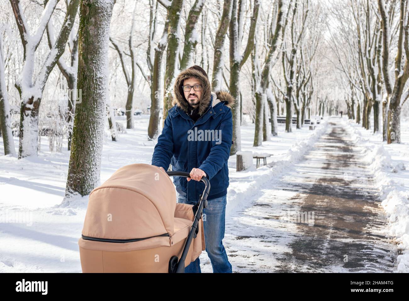 Caucasian man walking with buggy. Bearded father pushing baby stroller walking at winter park after blizzard. Spending time with infant in beautiful cold day. Stock Photo