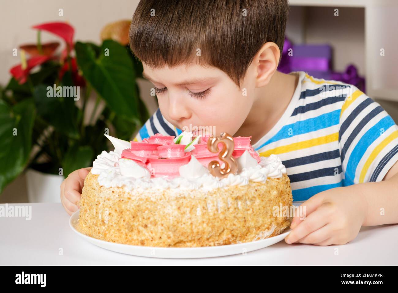 A child of 3 years old eats a birthday cake, licks the cream from the top sitting at the table Stock Photo