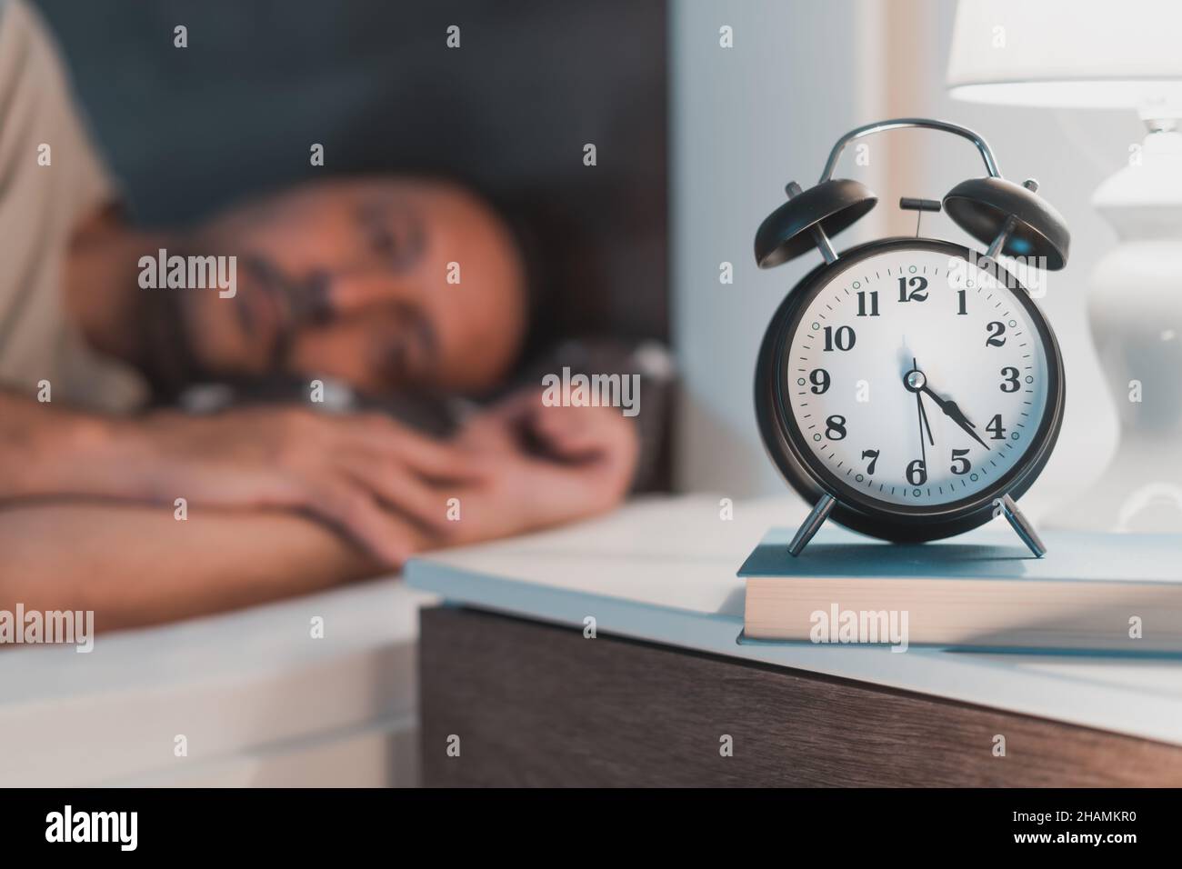 Sleepless man lying in bed and looking at alarm clock on bedside nightstand, selective focus Stock Photo