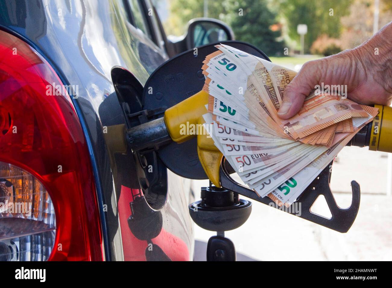 Illustration on the rise in pump prices. Someone filling a tank with petrol at a gas station. She’s holding a petrol pump and a bundle of 50 euro bank Stock Photo