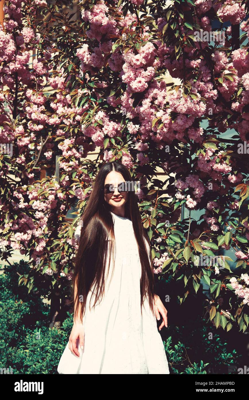 Beauty teen girl in white dress posing near sakura tree with lilac flowers. Spring park. Stock Photo