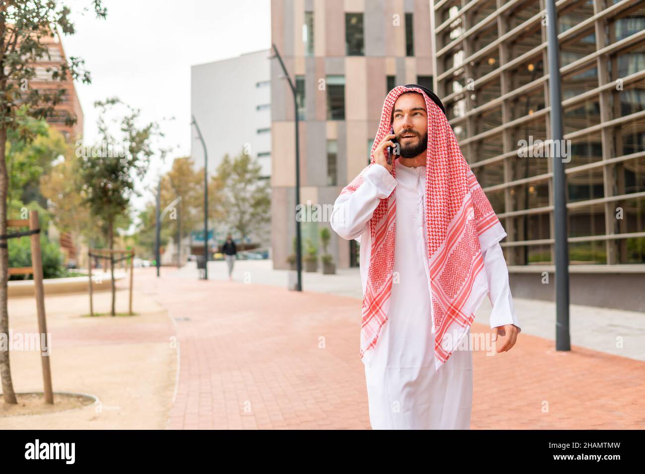 Arab businessman in traditional clothes making call on cellphone, talking on mobile phone with business partner while walking city street in Dubai, Saudi arab man using smartphone outdoors Stock Photo