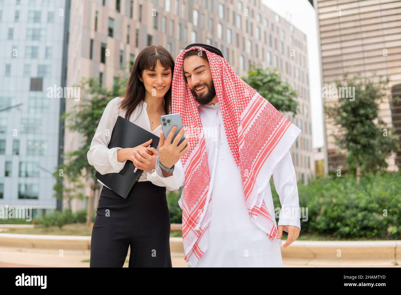 Smiling arab businessman in traditional wear showing video on smartphone to caucasian businesswoman at business meeting outdoors, saudi man and his female assistant looking at mobile phone outside Stock Photo