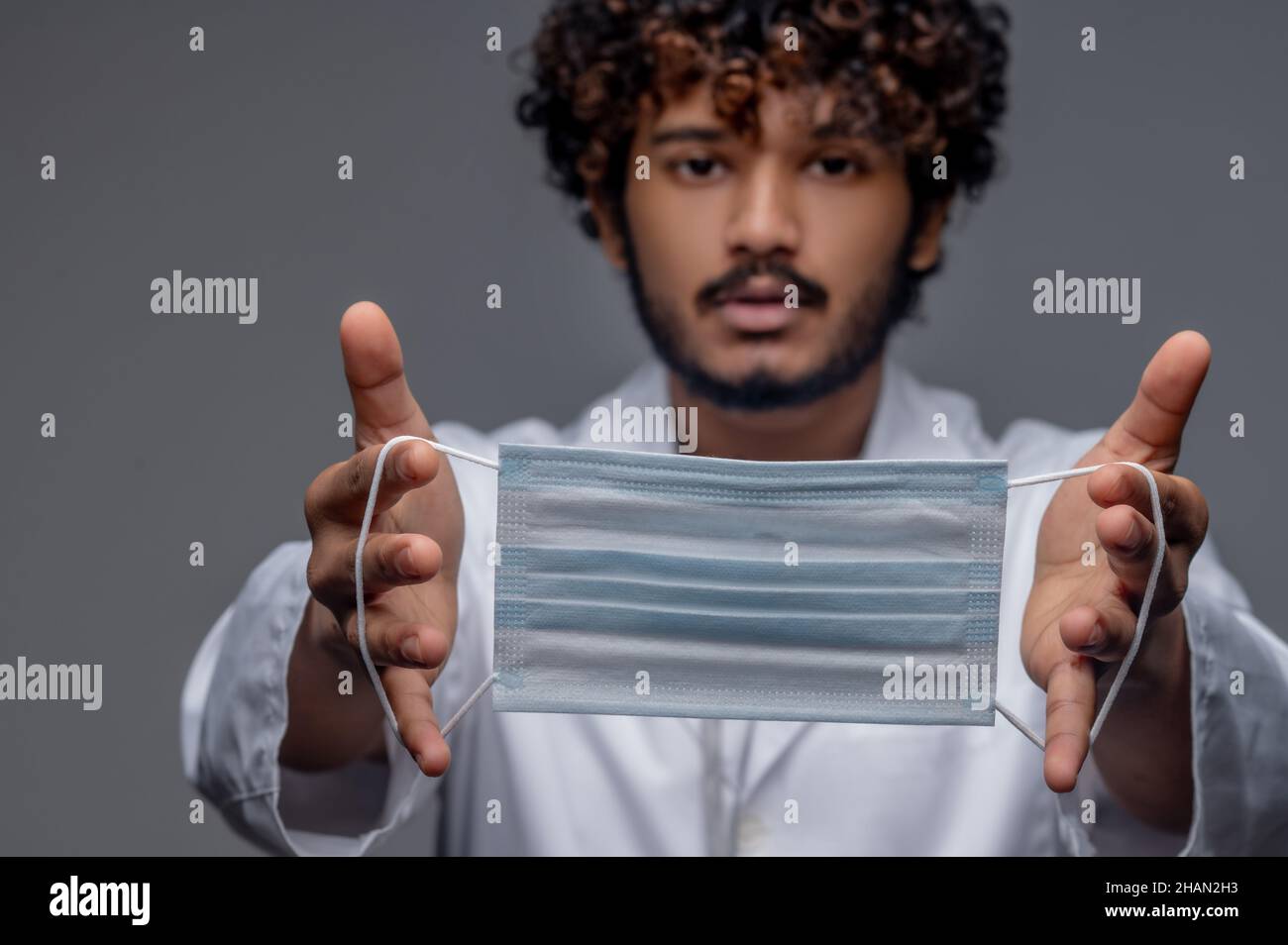 Bearded curly-haired physician holding a face covering before him Stock Photo