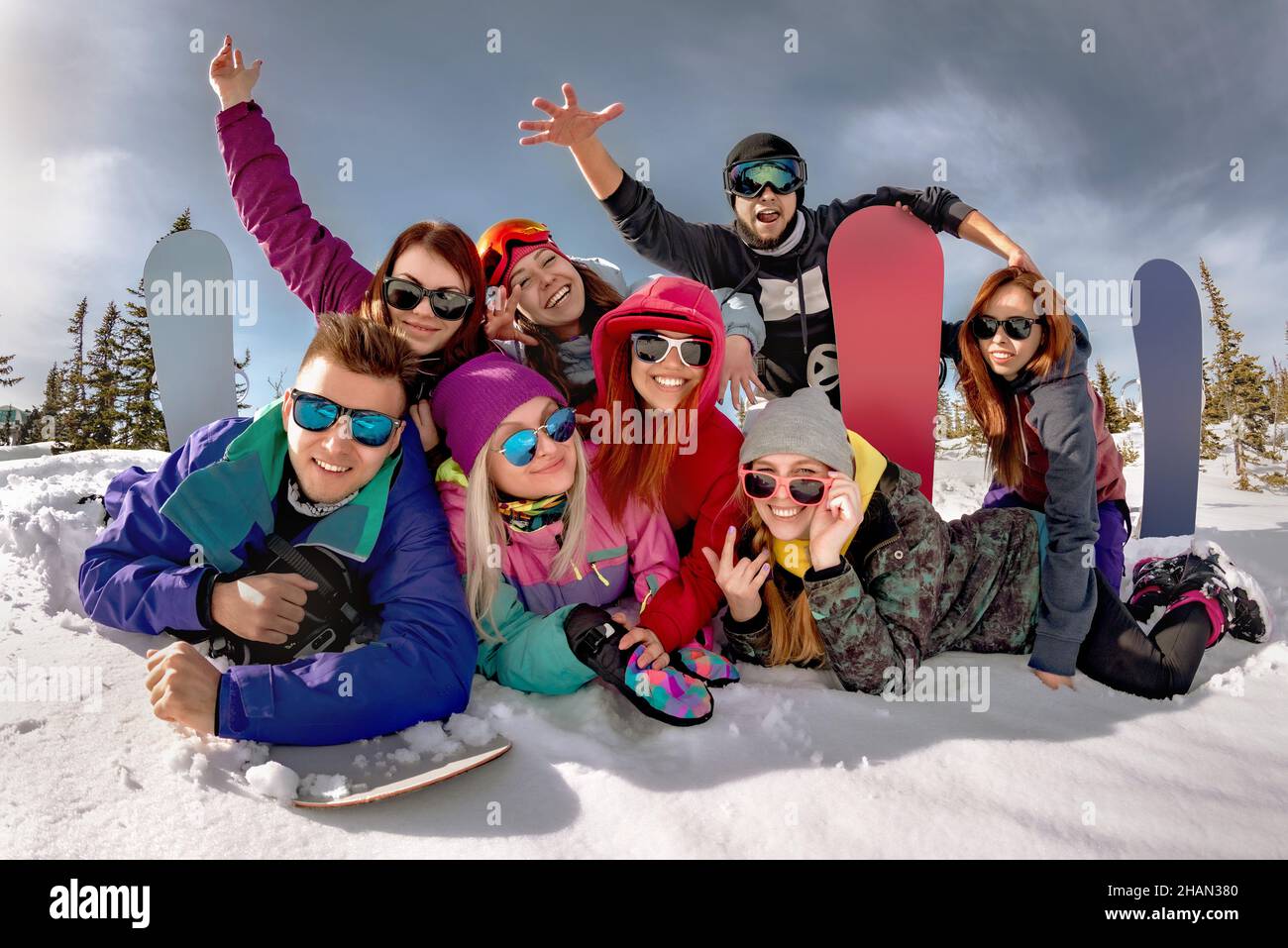 Group of happy friends, men and women sit in snow with ski and snowboards on winter weekend. Winter vacations concept. Stock Photo