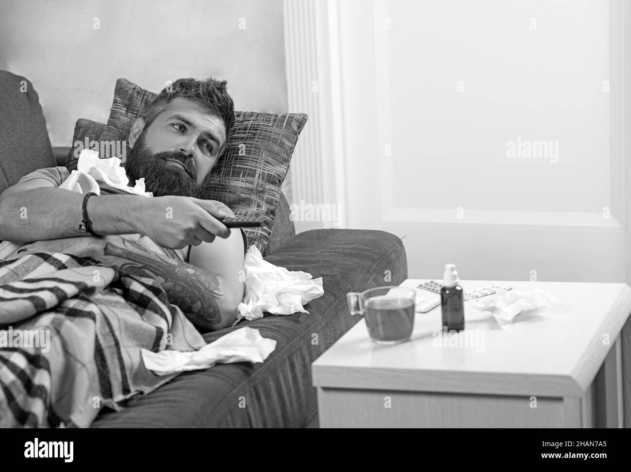 Bearded ill man lying in bed at home. Sick man lying on sofa with TV remote under a blanket at home in the living room. Stock Photo