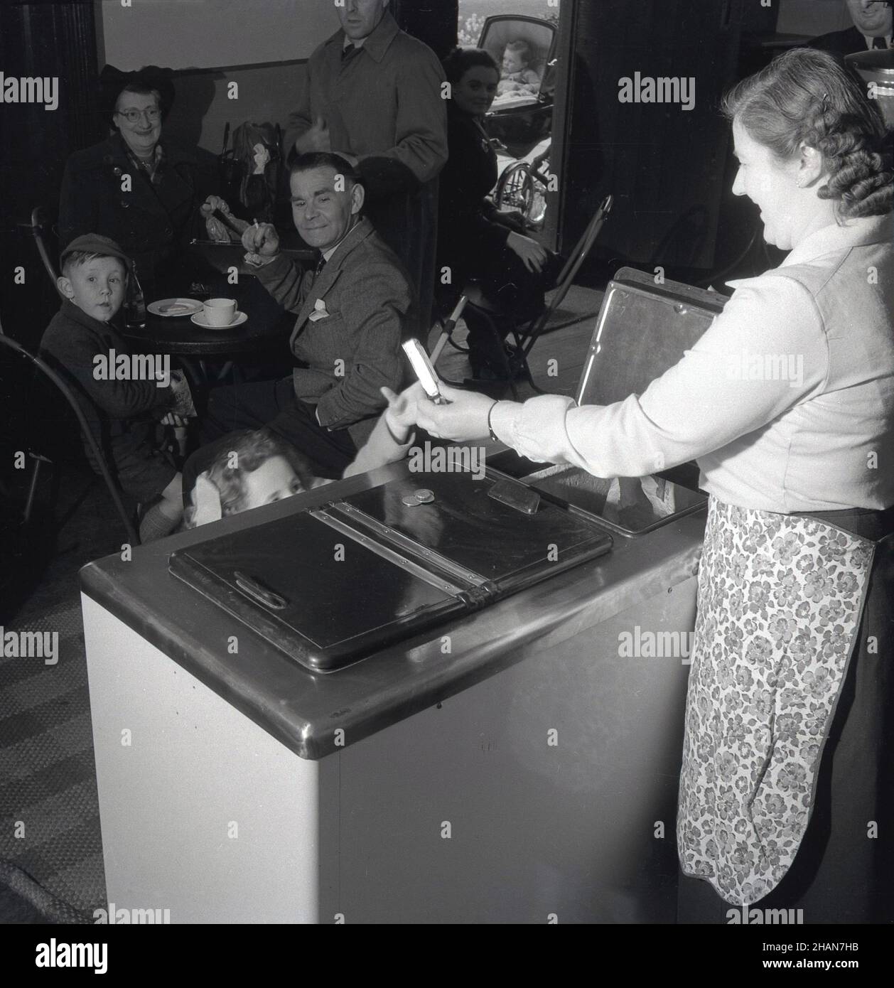 1950s, historical, inside a cafe, seated customers look on, as a little girl, having put some coins on the lid of the chest freezer, is given an ice cream wafer from the lady owner, London, England, UK. Stock Photo