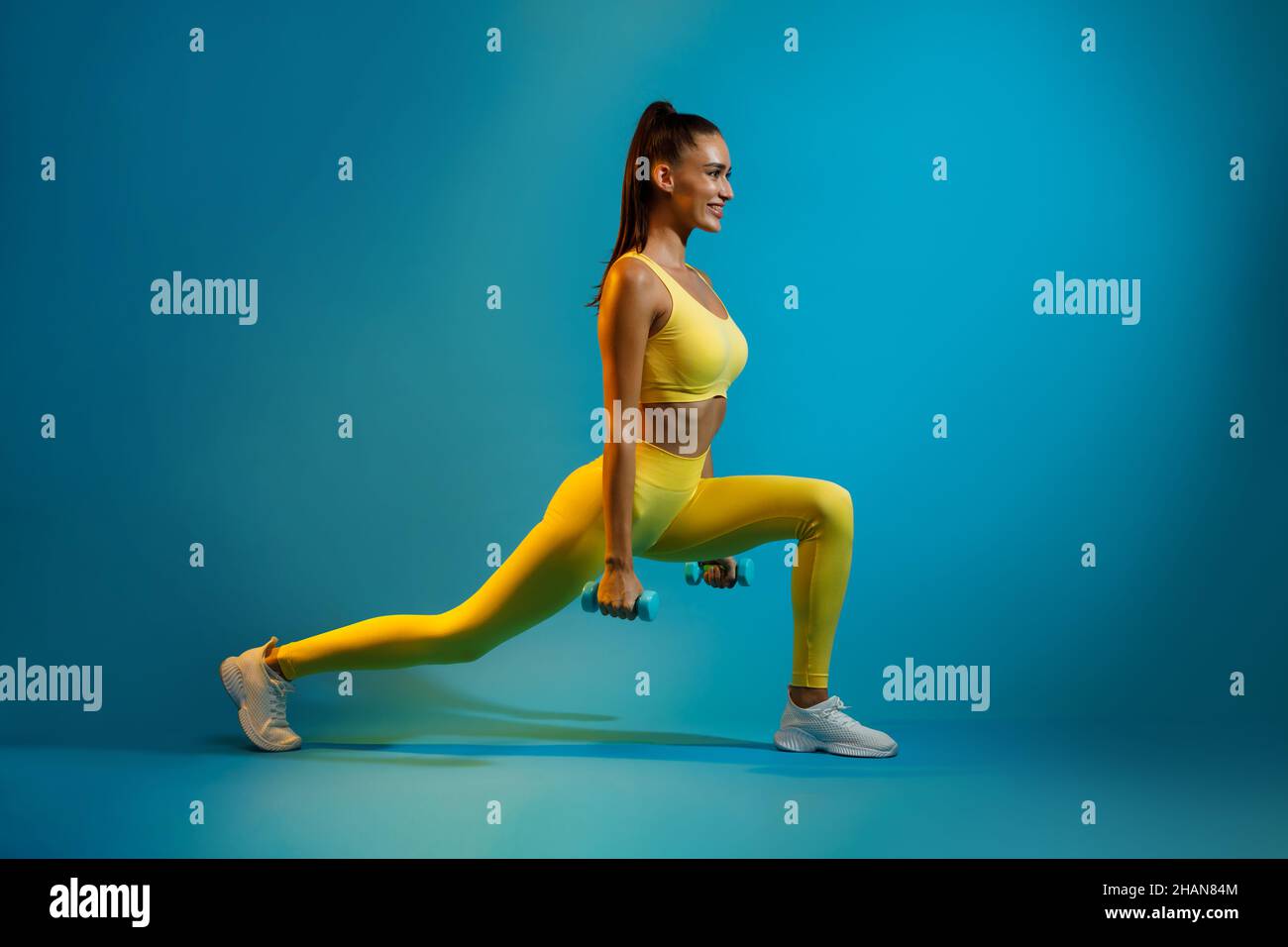 Sporty Female Doing Deep Lunge Exercise Holding Dumbbells, Blue Background Stock Photo