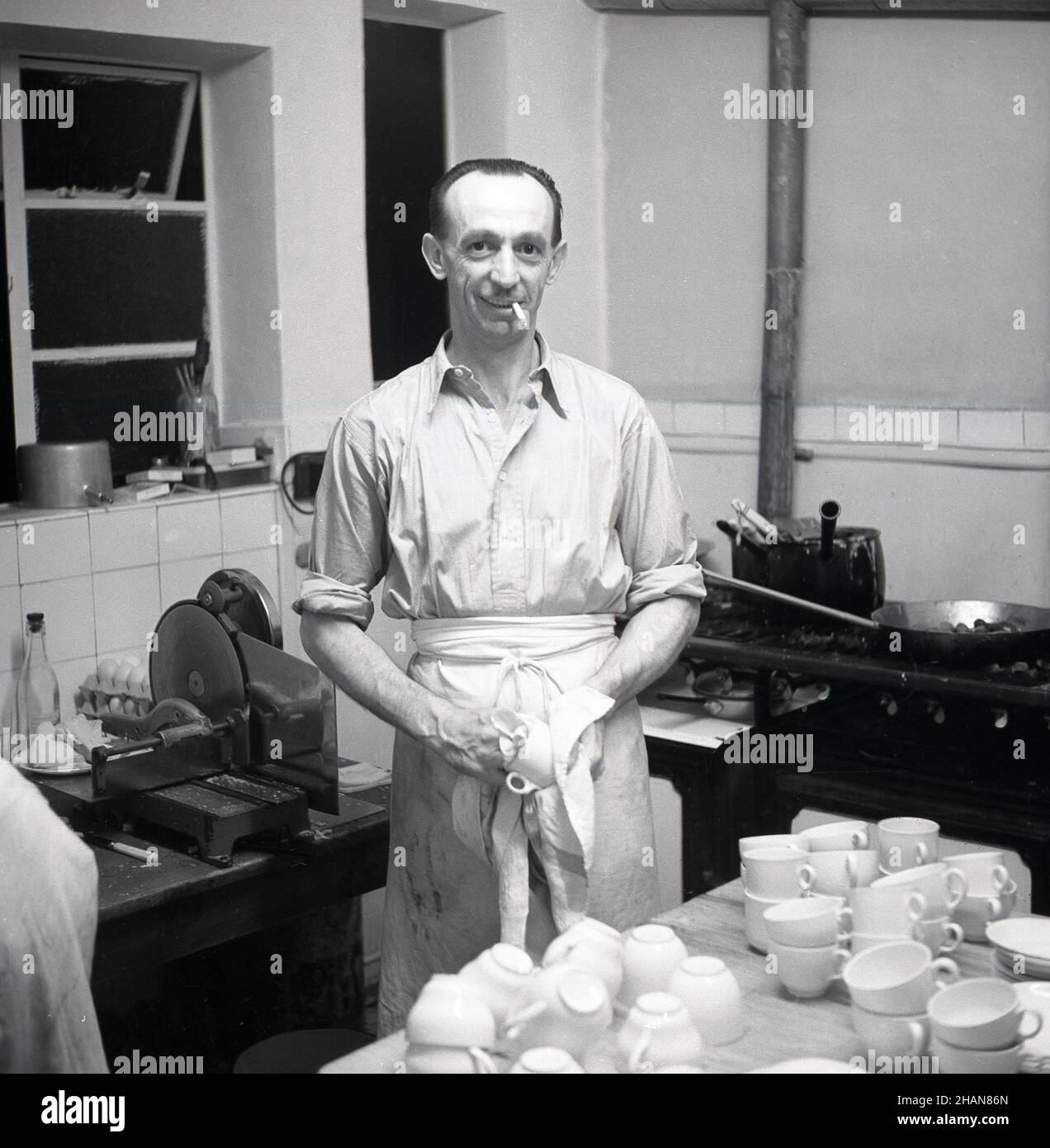1951, historical, a male cafe owner in his kitchen, wearing a waist or half apron, cigarette in mouth, wiping the cups dry with a tea towel, London, England, UK. Stock Photo