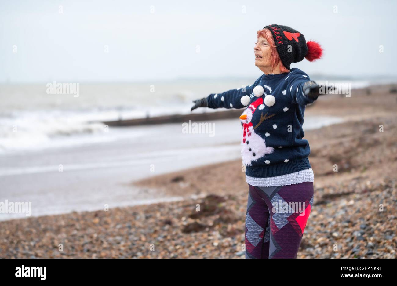 Elderly senior woman in her 80s on a beach exercising in Winter. She is wearing a Christmas jumper & hat while doing star jumps on the South Coast, UK Stock Photo