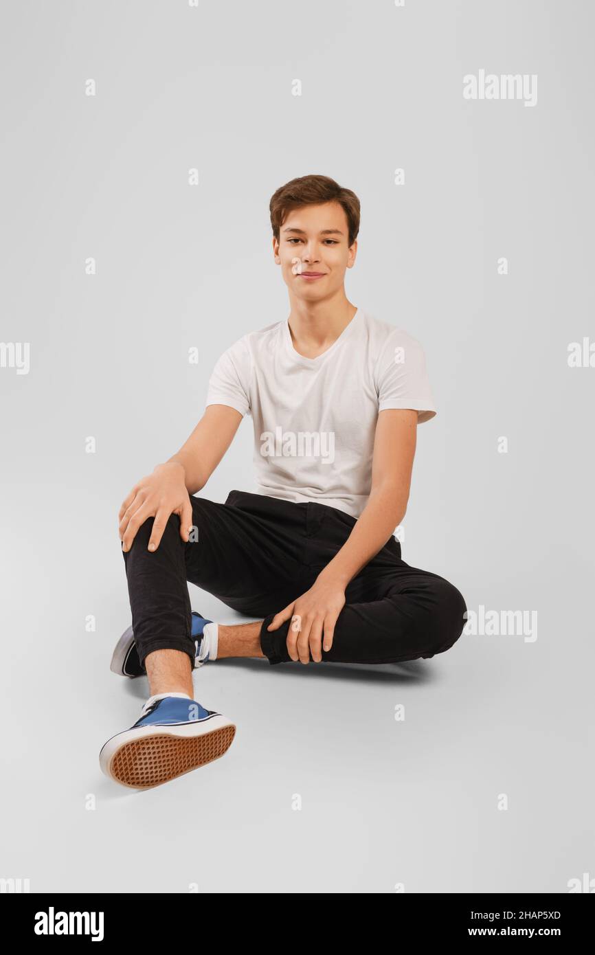 Handsome man in white t-shirt, black trousers and sneakers half laying  on the floor over grey background. Studio model tests. Stock Photo