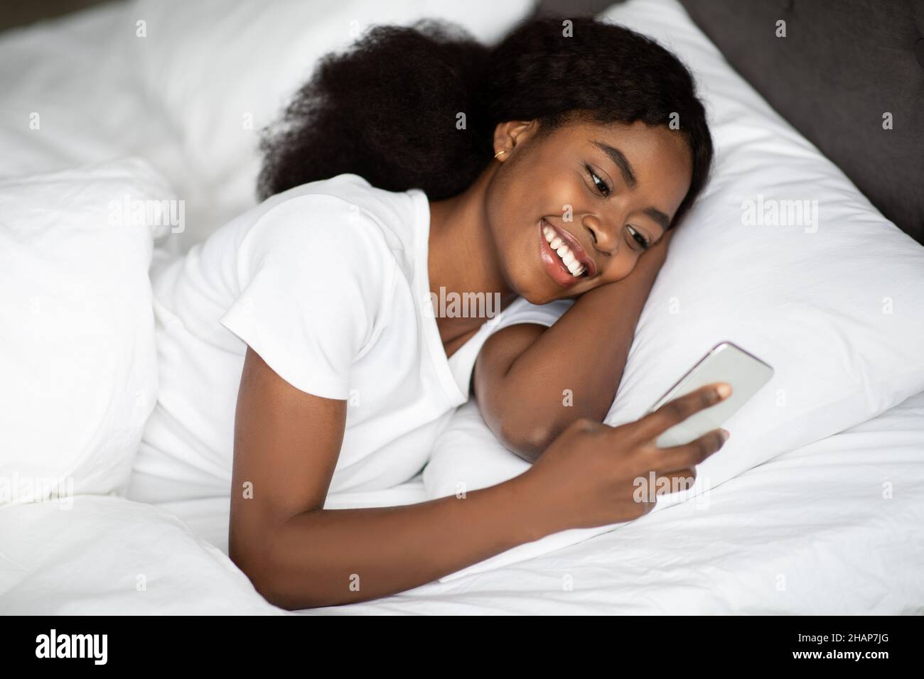 Positive young black lady laying in bed with smartphone Stock Photo