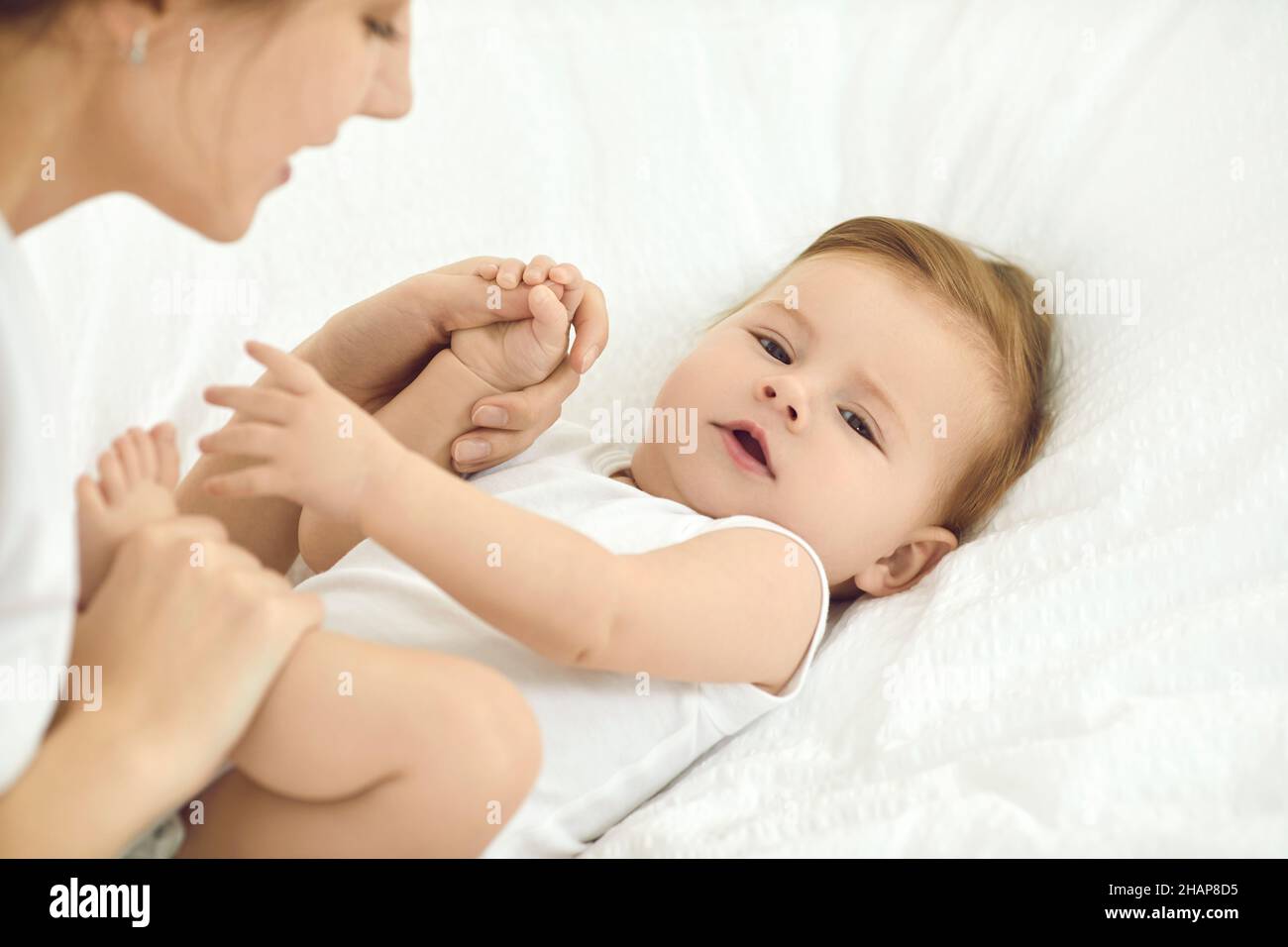 Portrait of adorable six month old baby girl lying on soft white bed and playing with mom. Stock Photo