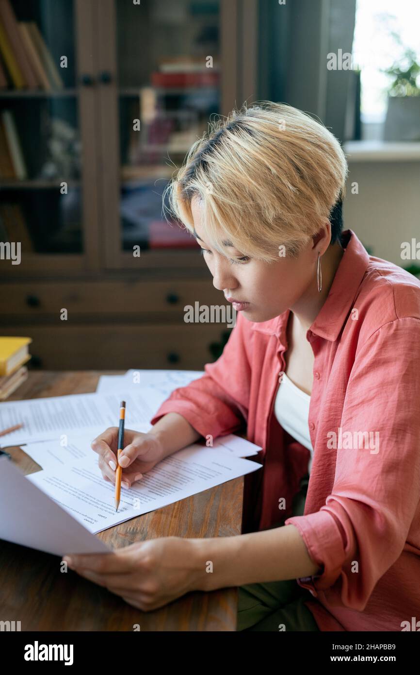 Young serious student or freelancer looking through papers and making notes while sitting by table in home environment Stock Photo