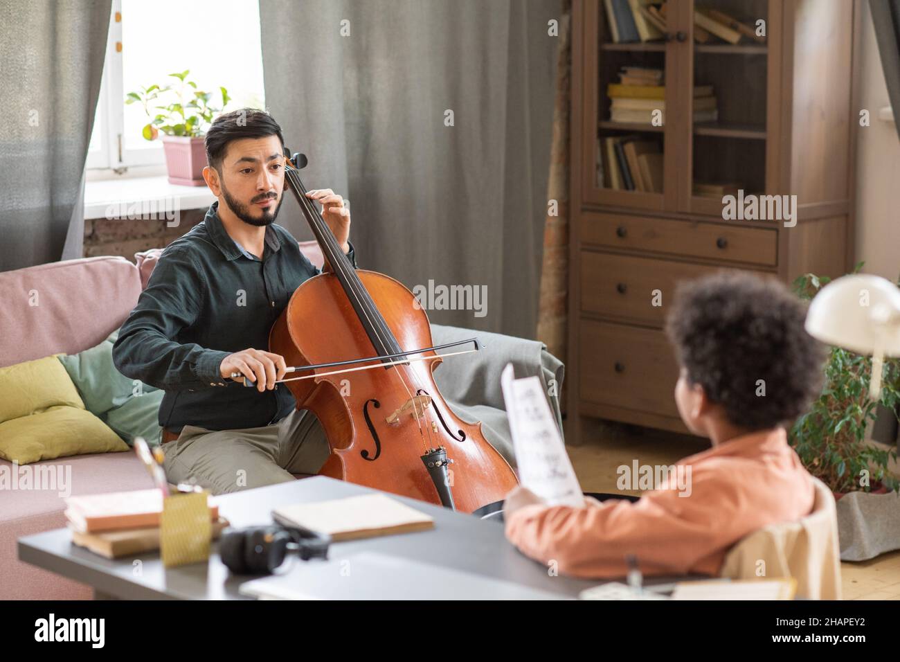 Young serious manle music teacher looking at schoolboy in front of him while playing cello during home lesson Stock Photo