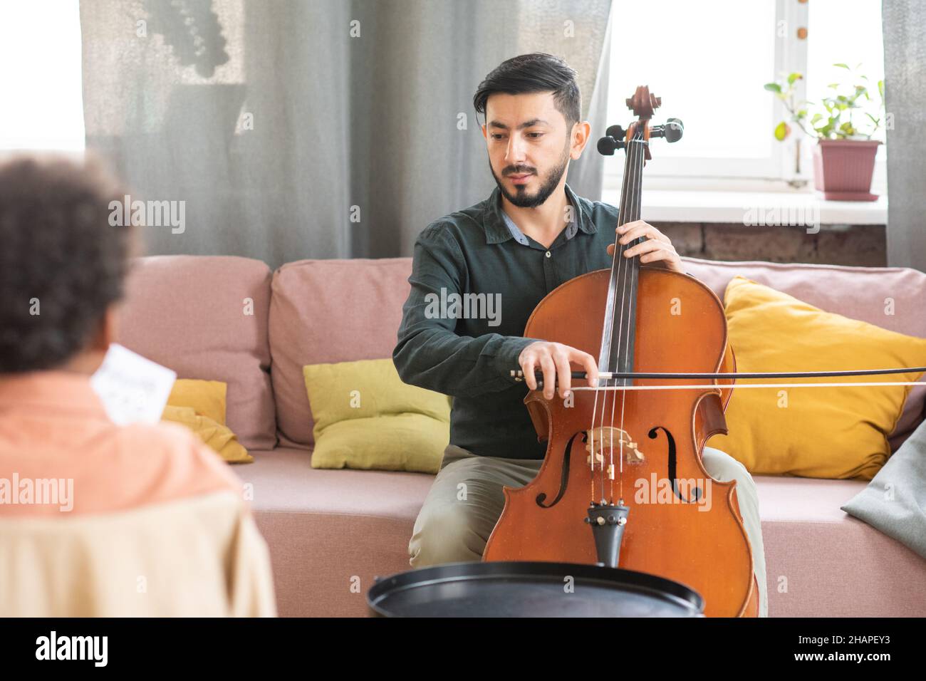 Young teacher of music sitting on couch and playing cello while teaching African schoolboy during home lesson Stock Photo