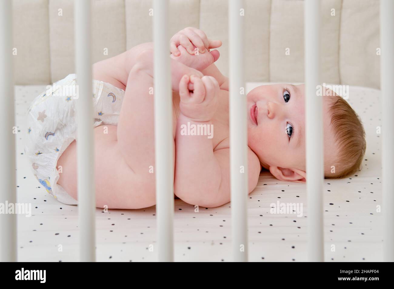 A happy infant baby with teething teeth in a diaper pulls his hand into his mouth. Funny child in a crib Stock Photo