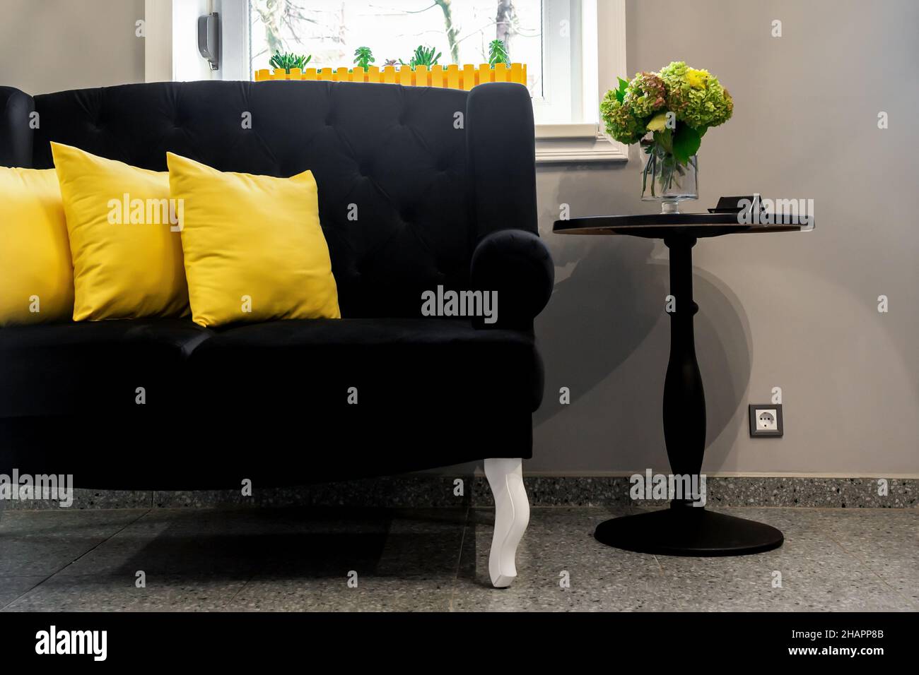 Interior of the beauty room. Black sofa with small yellow pillows and  coffee table on a gray wall background with a window Stock Photo - Alamy