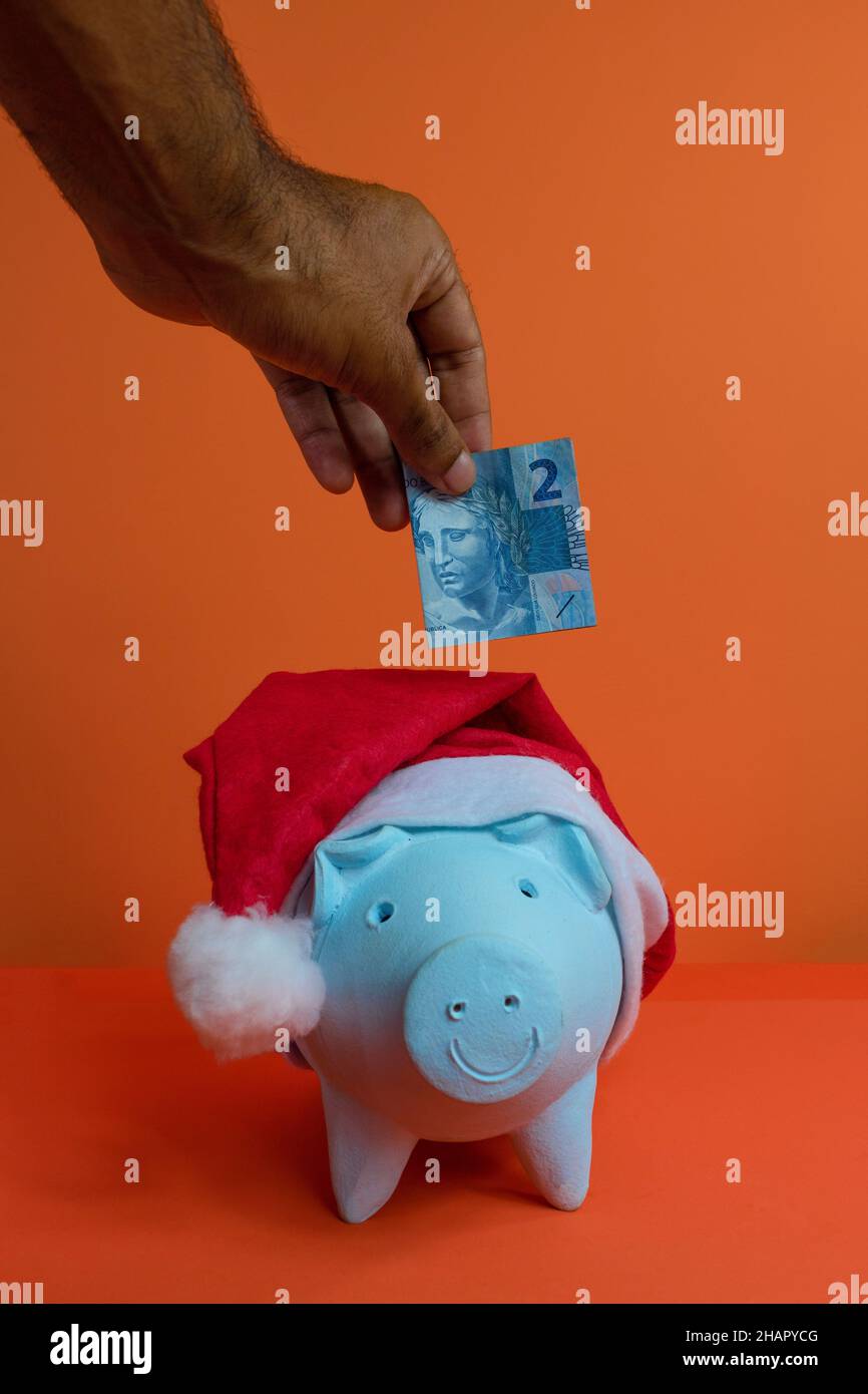 Hand putting real brazilian money in piggy bank with christmas santa claus hat isolated on orange background. Stock Photo