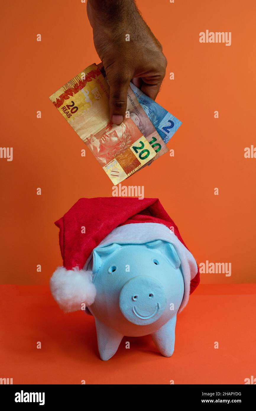 Hand putting real brazilian money in piggy bank with christmas santa claus hat isolated on orange background. Stock Photo