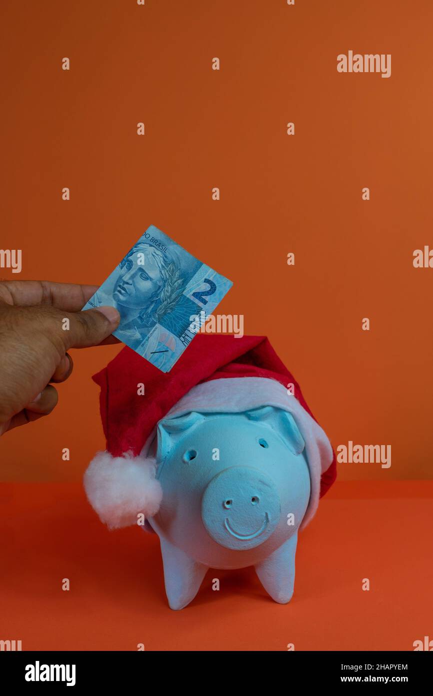 Hand putting real brazilian money in piggy bank with christmas santa claus hat isolated on orange background. Stock Photo