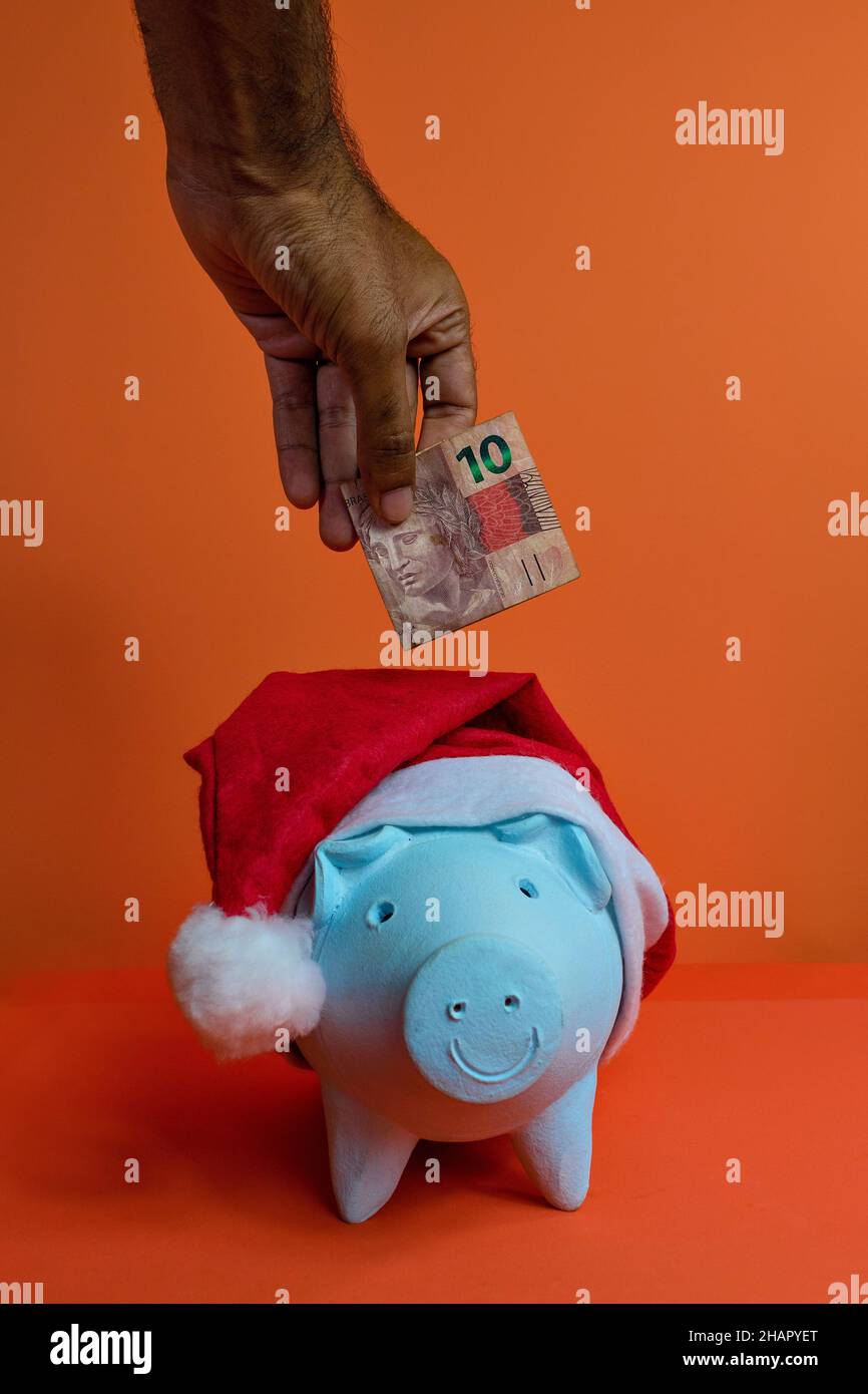 Hand putting real brazilian money in piggy bank with christmas santa claus hat isolated on orange background. Stock Photo