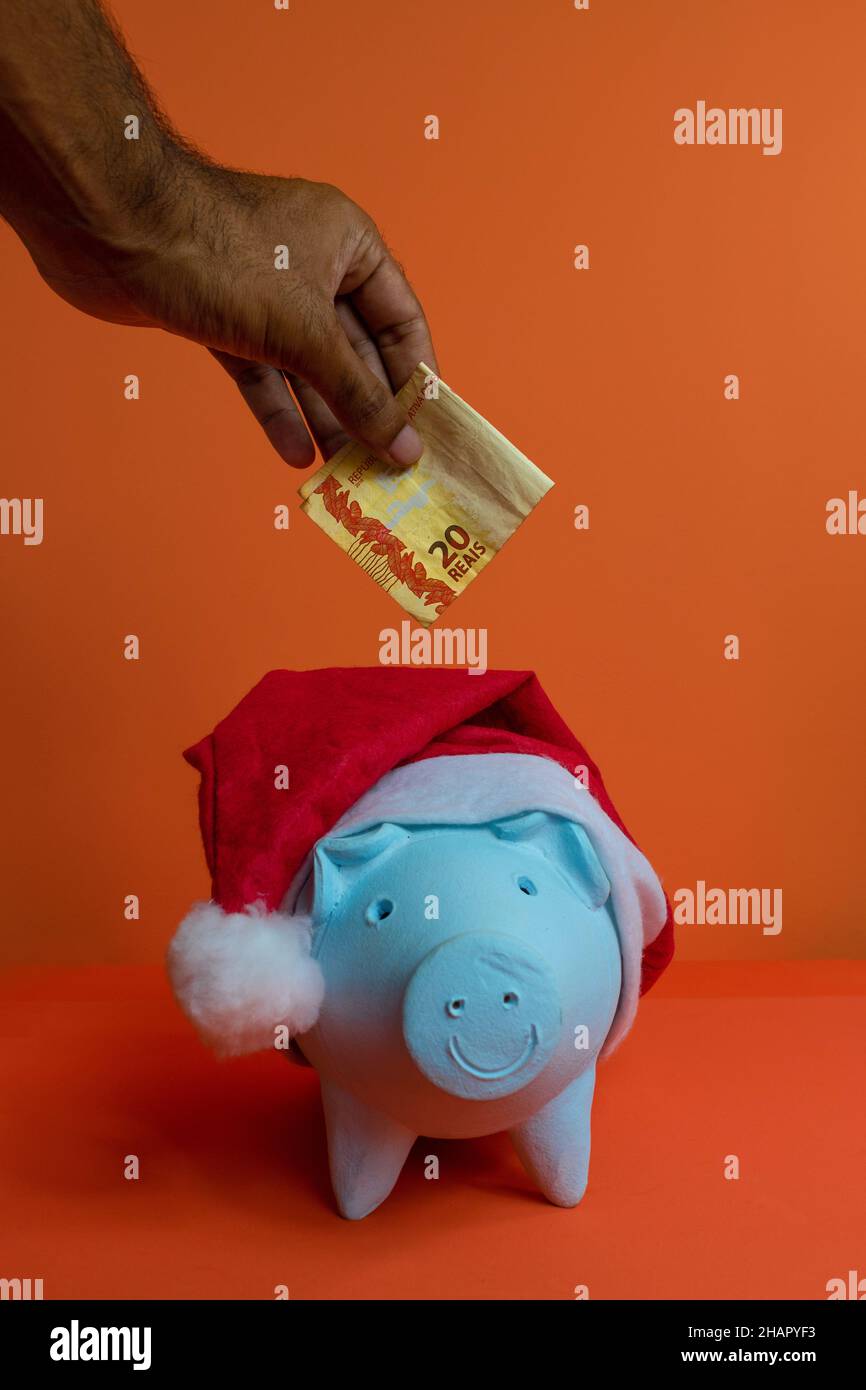 Hand putting real brazilian money in piggy bank with christmas santa claus hat isolated on orange background. Stock Photo