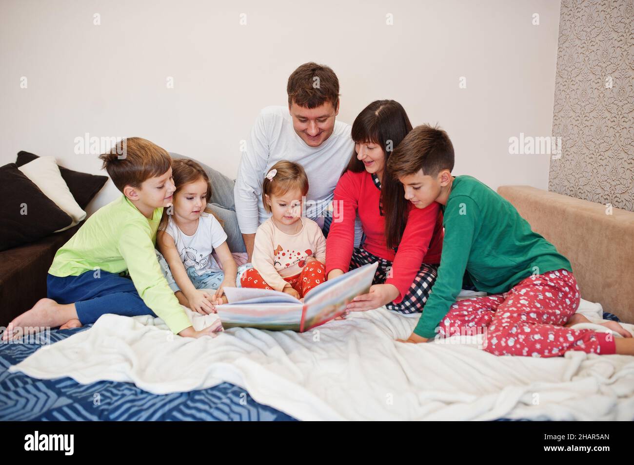 Happy big family is having fun together in bedroom. Large family morning concept. Four kids with parents wear pajamas read book in bed at home. Stock Photo