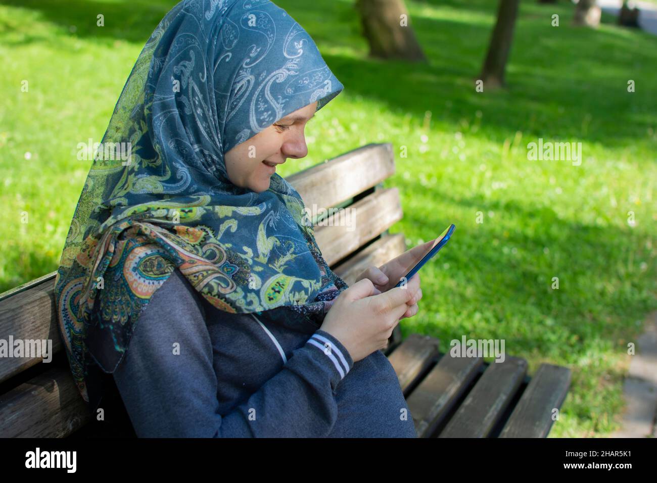 Profile view of the muslim pregnant woman smiling while using mobile phone and sitting alone in the park Stock Photo